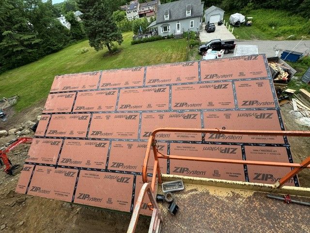 Roof under construction with orange ZIP sheathing panels, lumber, and a house visible in the background.