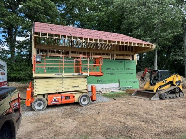 Construction site with house framing, orange lift, and yellow skid steer on a dirt lot