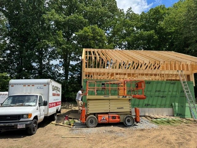 Work truck and worker beside a house under construction, with exposed roof framing in a wooded yard.