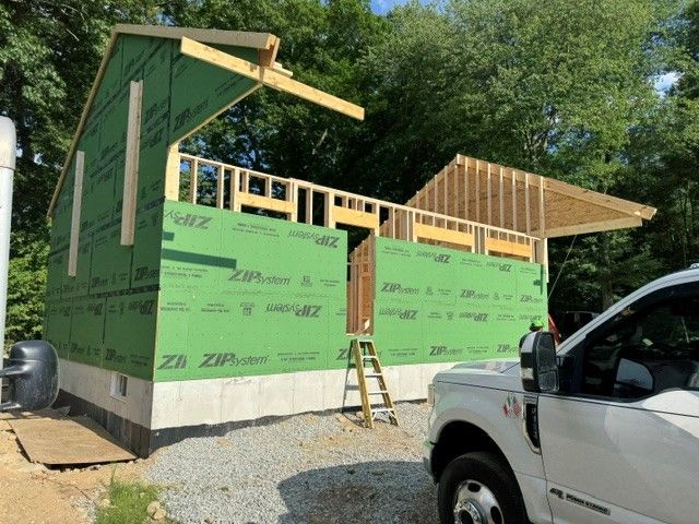 Partially built house with green sheathing, exposed wood framing, and a ladder beside it under a blue sky