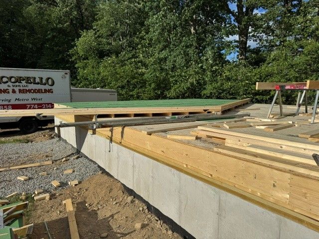 Construction site with wooden framing and concrete foundation, surrounded by trees and gravel.
