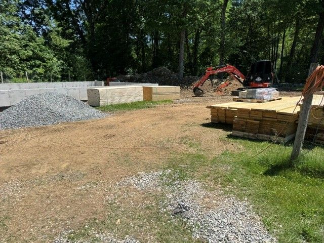 Construction site with gravel piles, stacked materials, and a red excavator in a wooded area