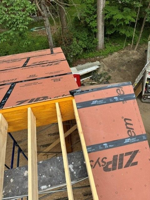 Roof construction with orange Zip System sheathing on a steep roof beside trees and dirt ground
