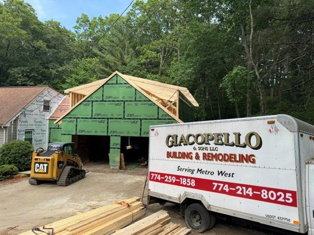 House under construction with green sheathing, a CAT loader, and a roofing truck in front of a wooded area