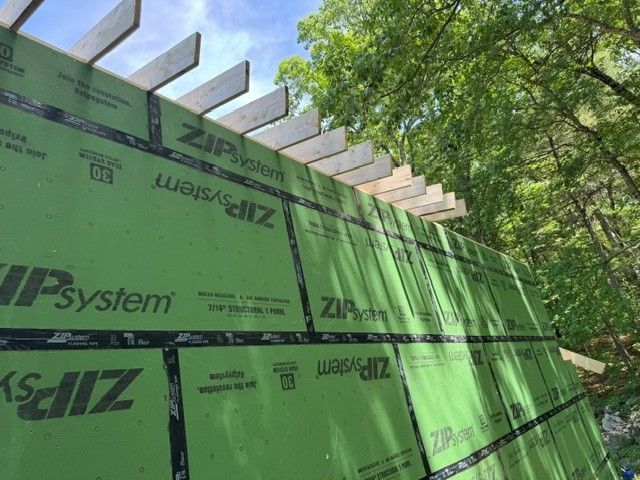 Green ZIP System sheathing on a framed house wall under construction, with roof rafters and trees overhead