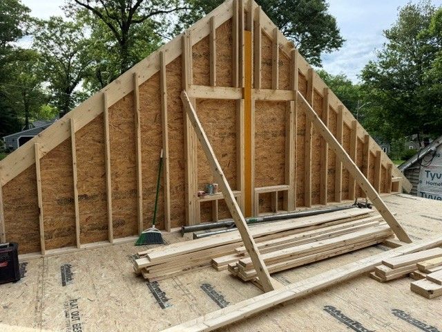 Wood-framed house under construction with stacked lumber and trees in the background