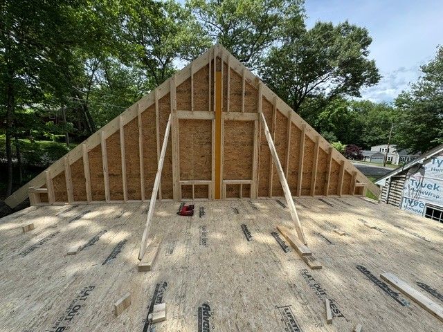 Framed gabled roof under construction on a wood deck, with trees in the background