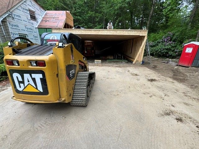 Yellow CAT excavator on a dirt driveway beside a shed, with a red portable toilet in the background.