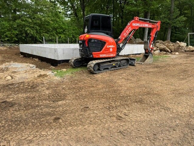Orange mini excavator beside a concrete foundation slab on a dirt lot in a wooded area