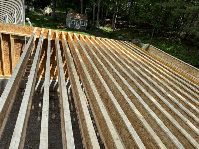Wooden roof framing under construction on a house, with exposed rafters and a wooded background.