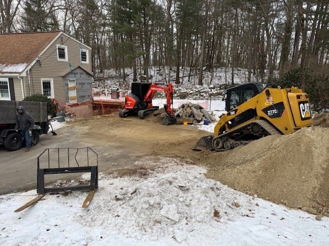 Construction site with a backhoe, skid steer, and dump truck. Dirt and snow on the ground near a house and trees.