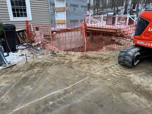 Construction site: Excavator beside a hole. House and partially built deck visible; orange safety fencing.