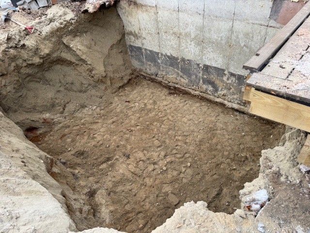 Excavated foundation pit. Soil and foundation walls visible. Wooden boards are near the top right.