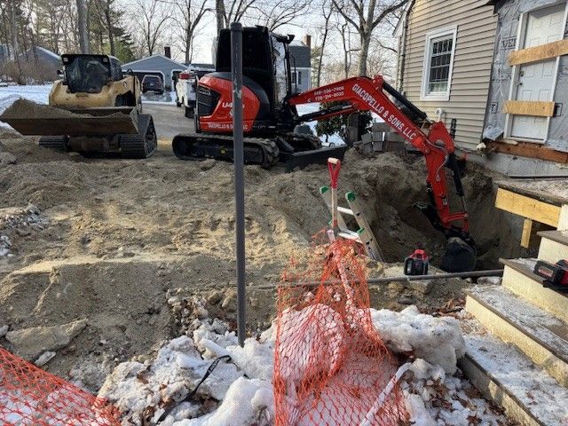 An excavator digs near a house, orange safety netting in the foreground, snow on the ground.