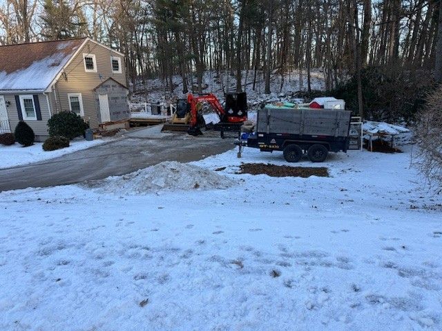 Snowy scene: Excavator on residential property, trailer, house in background. Construction in progress.