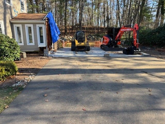 A blacktop driveway leads to a concrete pad with construction equipment: a skid steer and excavator near a building.