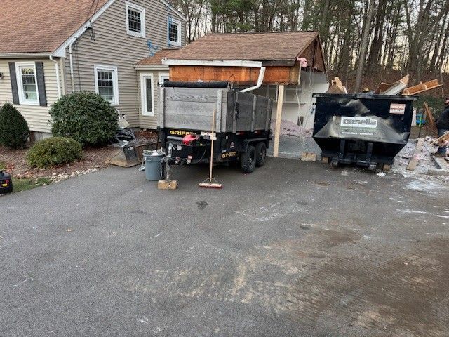 Dump trucks and a dumpster sit in a driveway next to a house under construction.