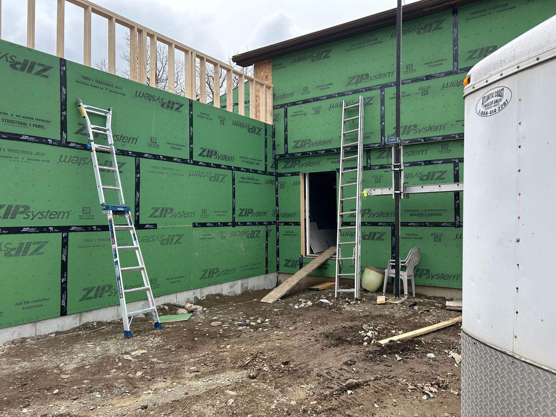Exterior of a building under construction. Green panels on the walls. Ladders, dirt ground, and a trailer present.