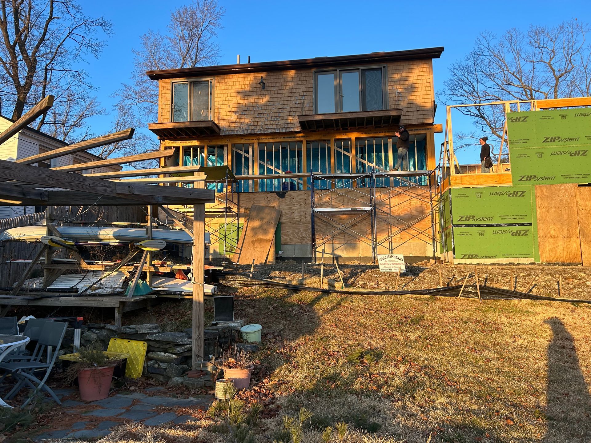 Two-story house with balcony and wooden siding under construction. Person on right-side scaffolding. Sunny outdoor setting.