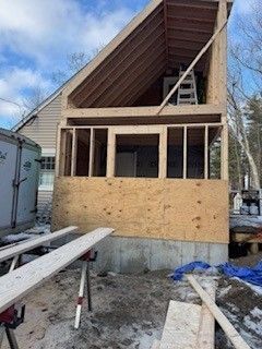 Construction of a house addition. Wooden framing with plywood siding. A-frame roof. Outdoor setting.