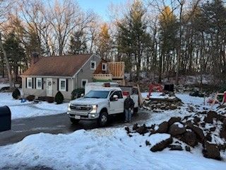 A white pickup truck parked in a snowy driveway with a person standing next to it. A house with construction is in the background.