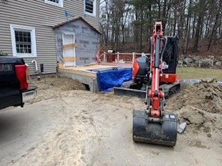 Mini excavator digging near a house, possibly for deck construction, with a pickup truck nearby.
