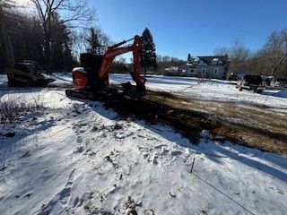 An excavator digs a trench in a snow-covered field, with a house and trees in the background under a blue sky.