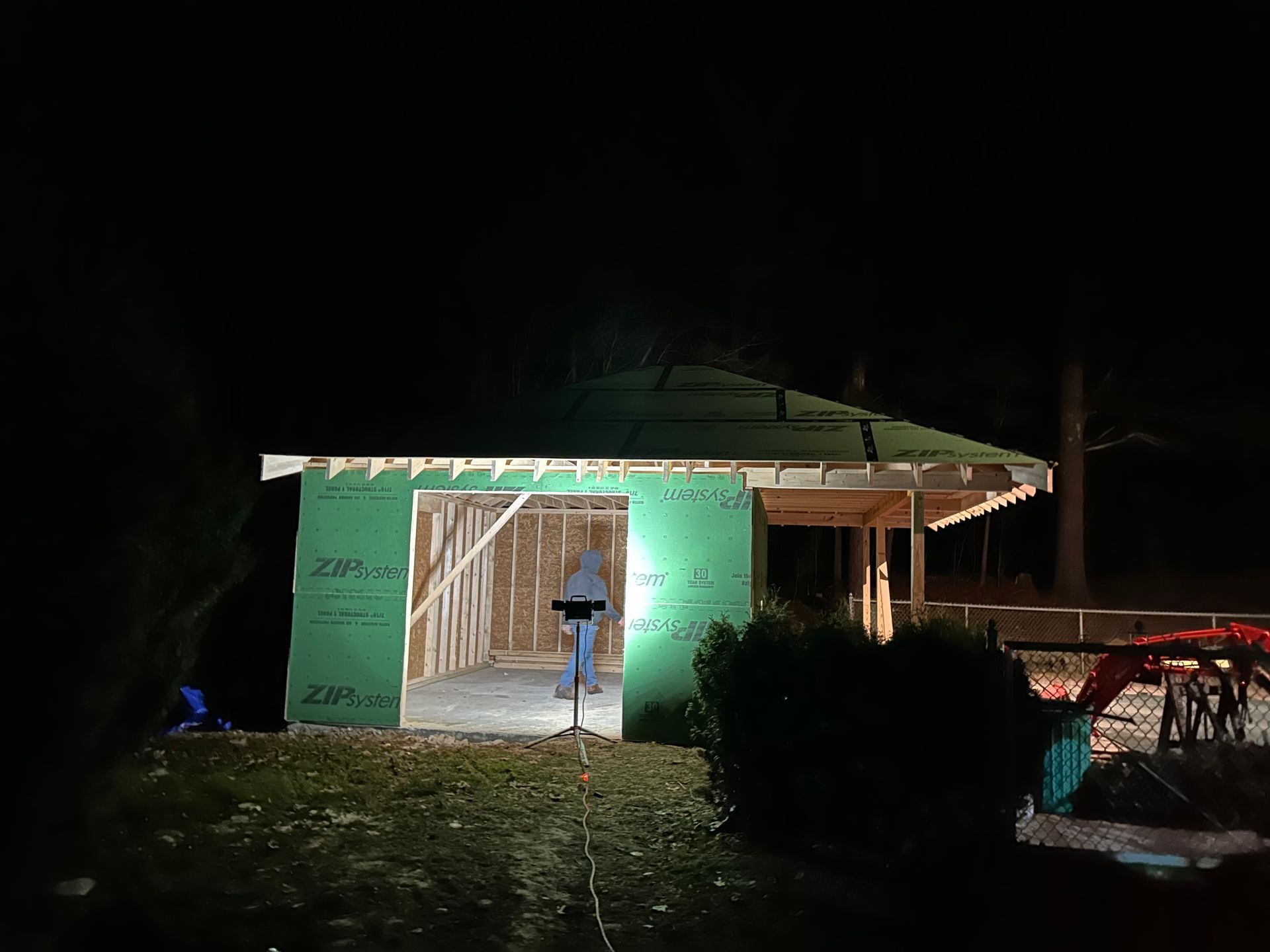 Garage under construction at night, green siding, framing visible, person working with light.