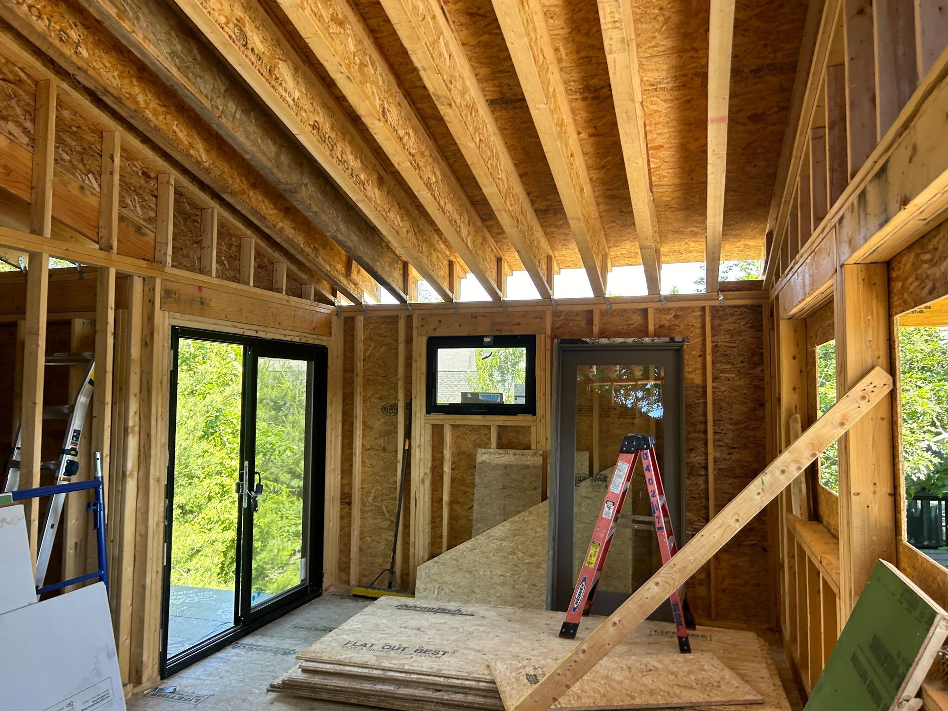 Interior framing of a building under construction, with exposed wooden beams, windows, and a sliding glass door.
