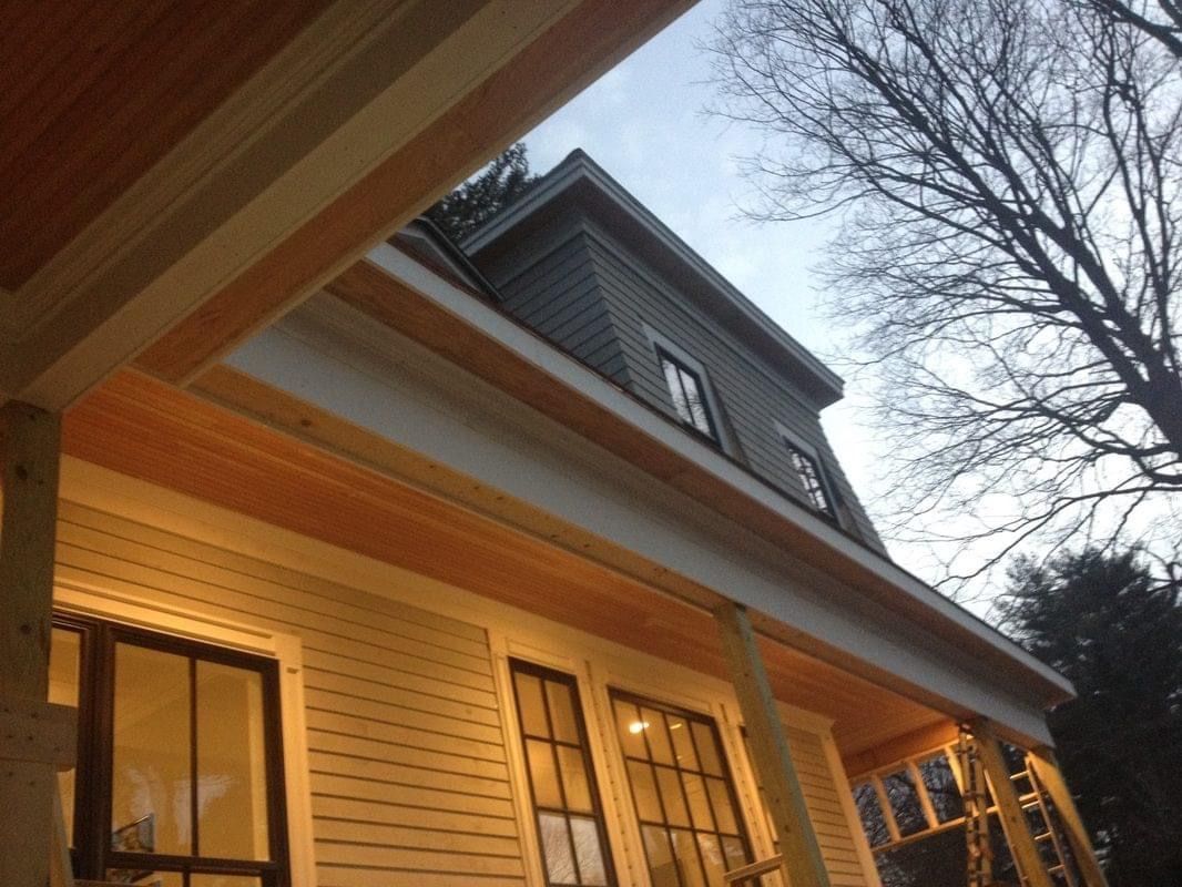 View of a home's porch and dormer; light wood and white trim, light siding, windows, and a cloudy sky.