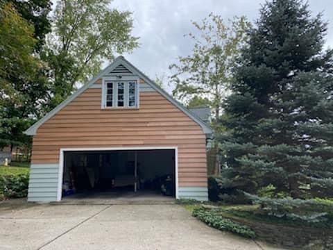 Garage with open door, brown siding, and small window on top. Tall evergreen tree to the right.