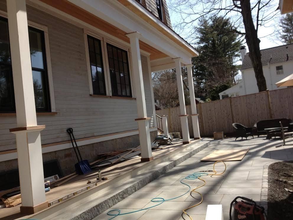 A light gray house with a porch under construction, paved patio, and wooden fence.