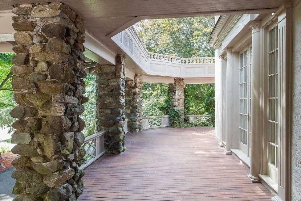 Covered porch with stone columns and wooden floor, overlooking a garden.