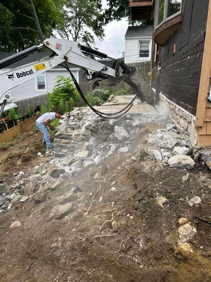 Bobcat demolishes stone walkway near a house; worker nearby. Debris and dust fill the air.