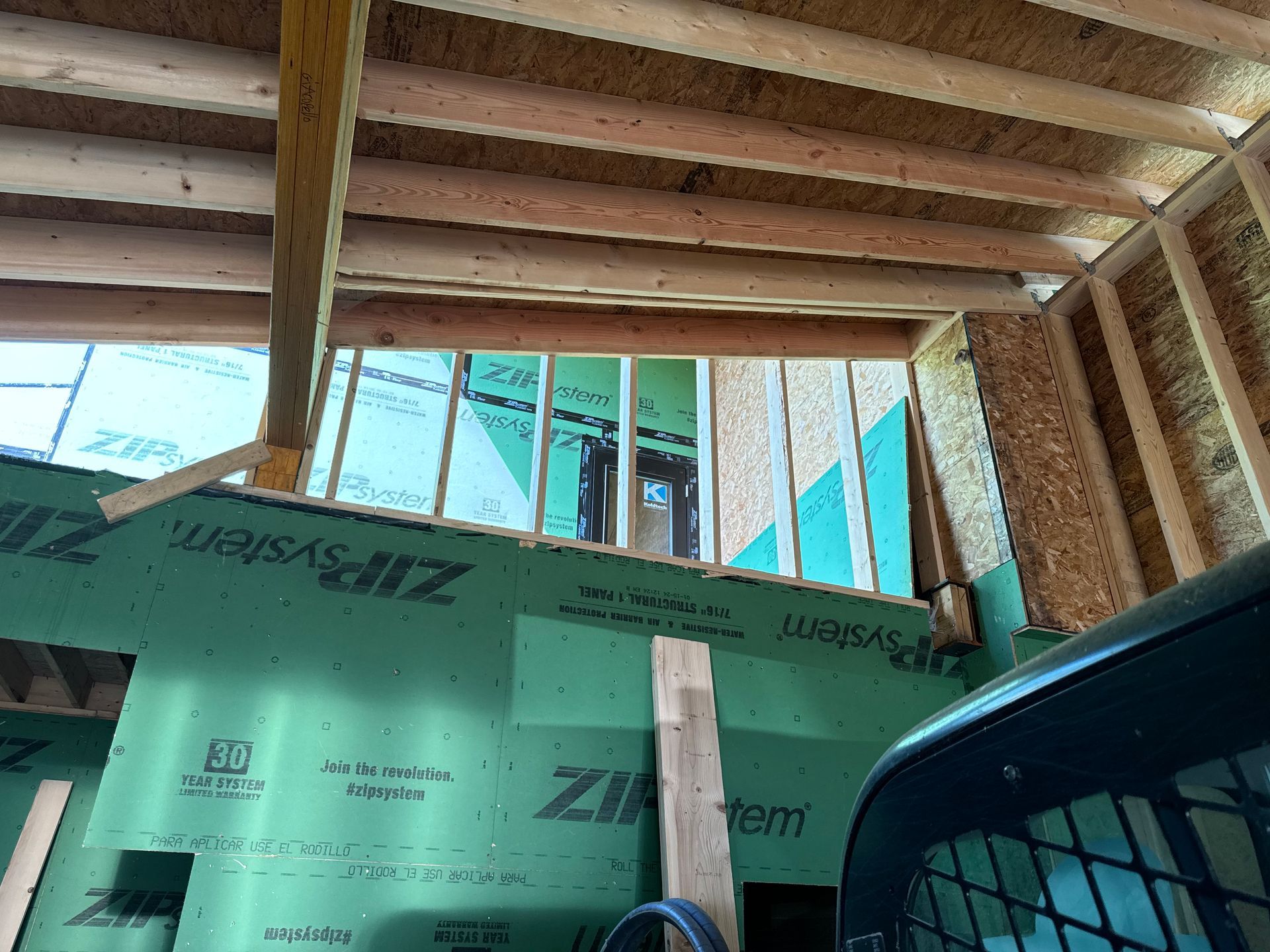 Interior view of a building under construction, showing wooden framing, green sheathing, and a partially built window.