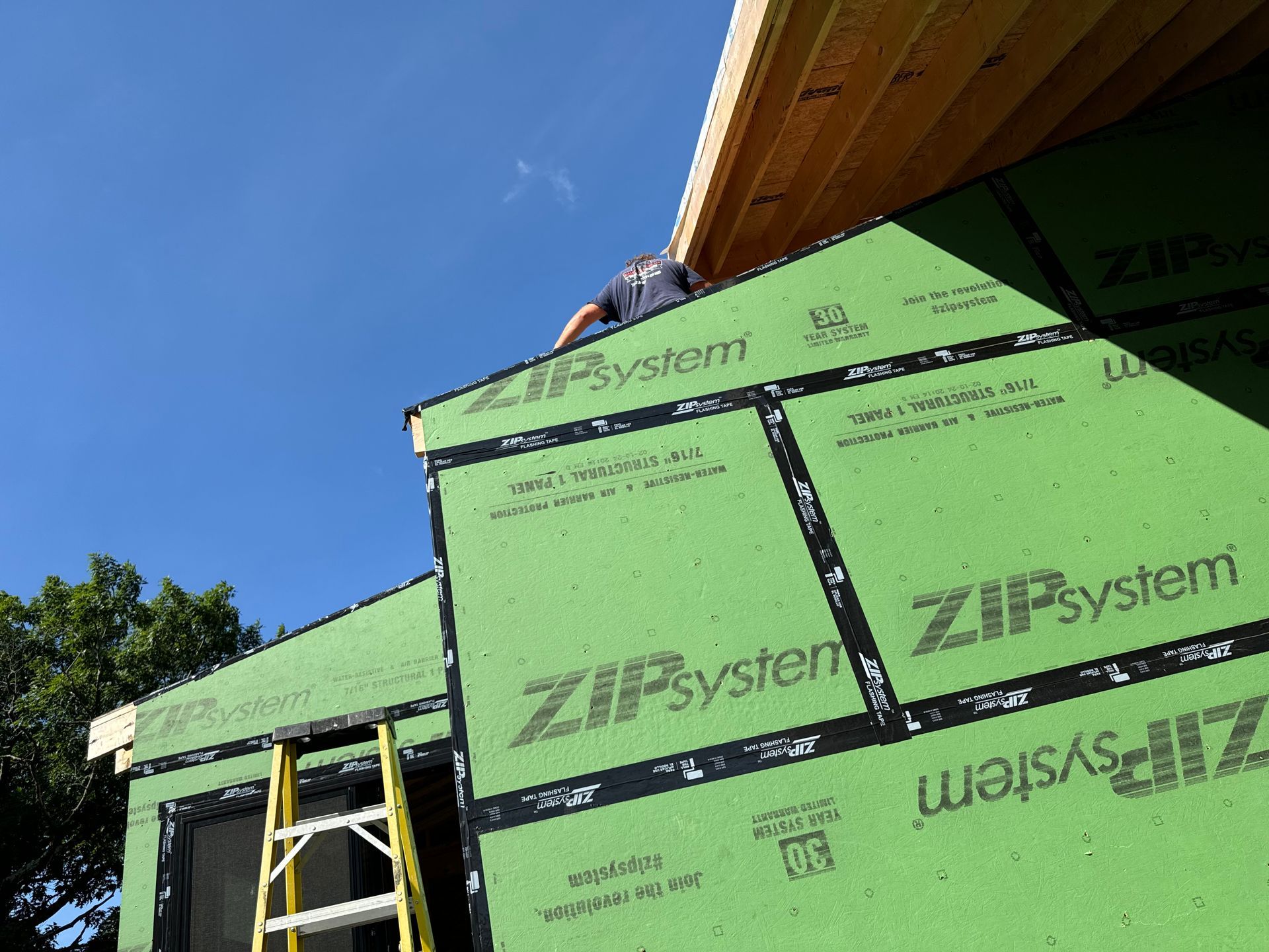 Construction worker installing green Zip System sheathing on a building under a blue sky, ladder visible.