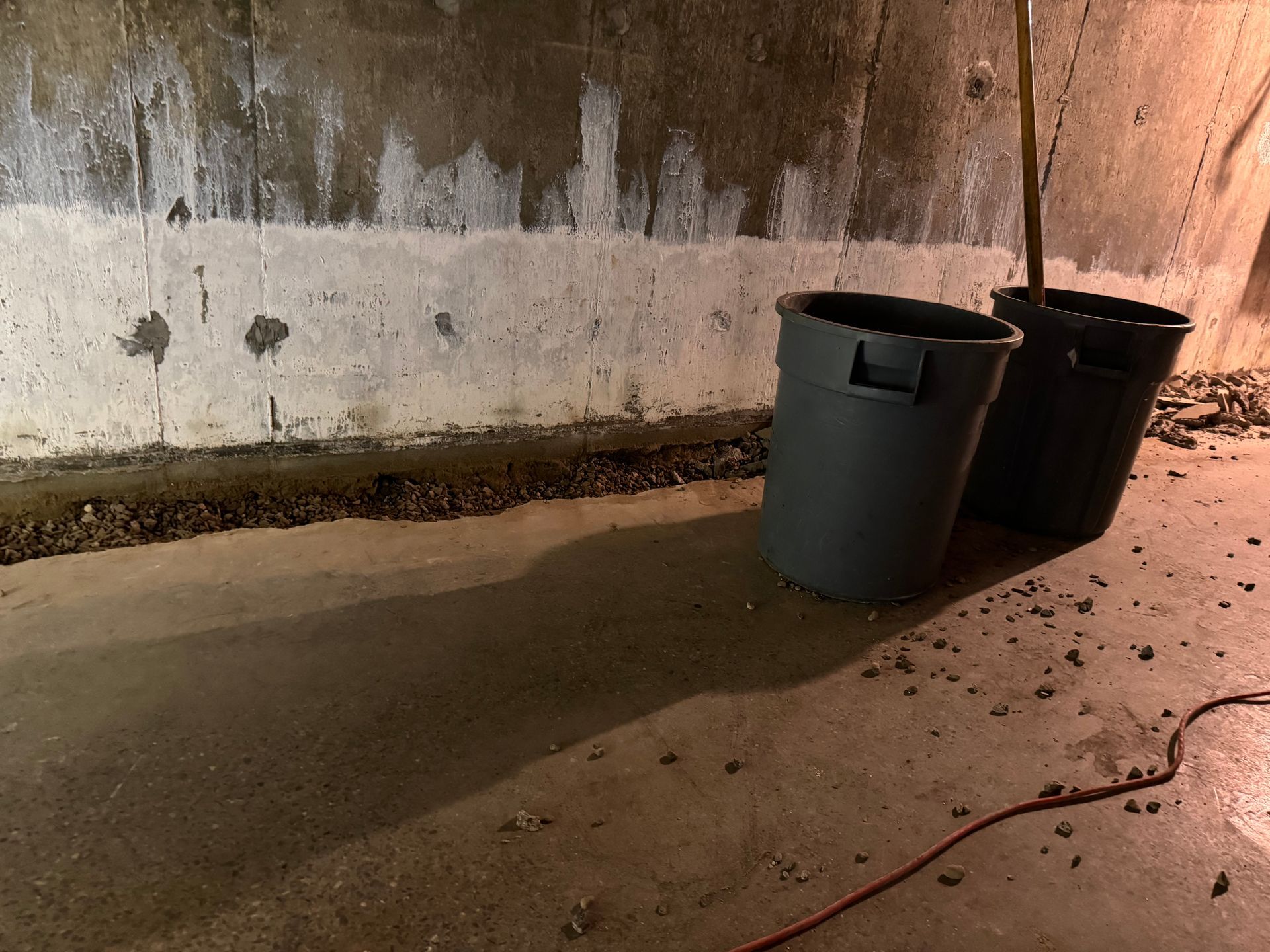 Two gray trash cans next to a concrete wall in a dimly lit basement.