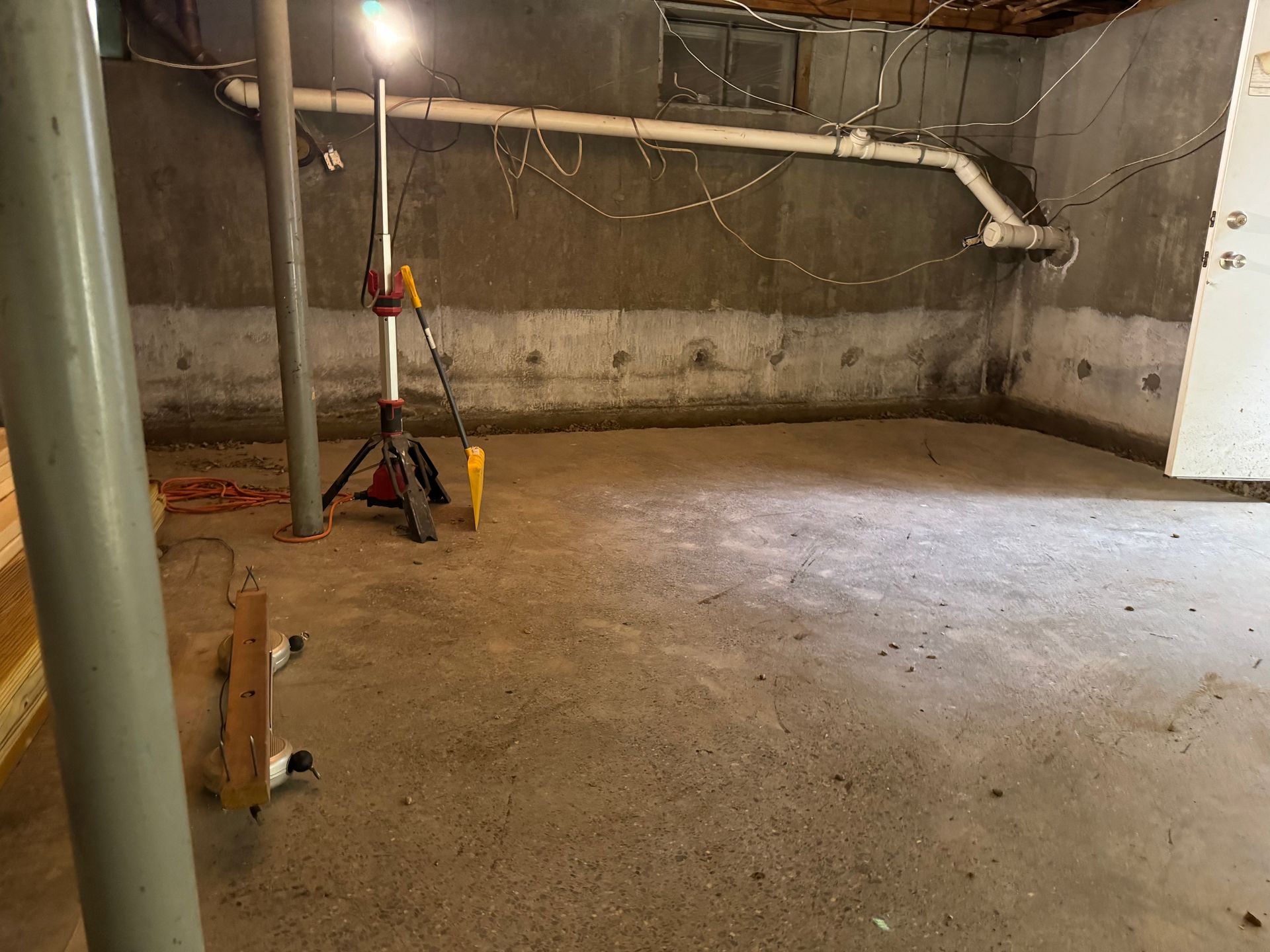A concrete basement with exposed pipes, wiring, and a work light.