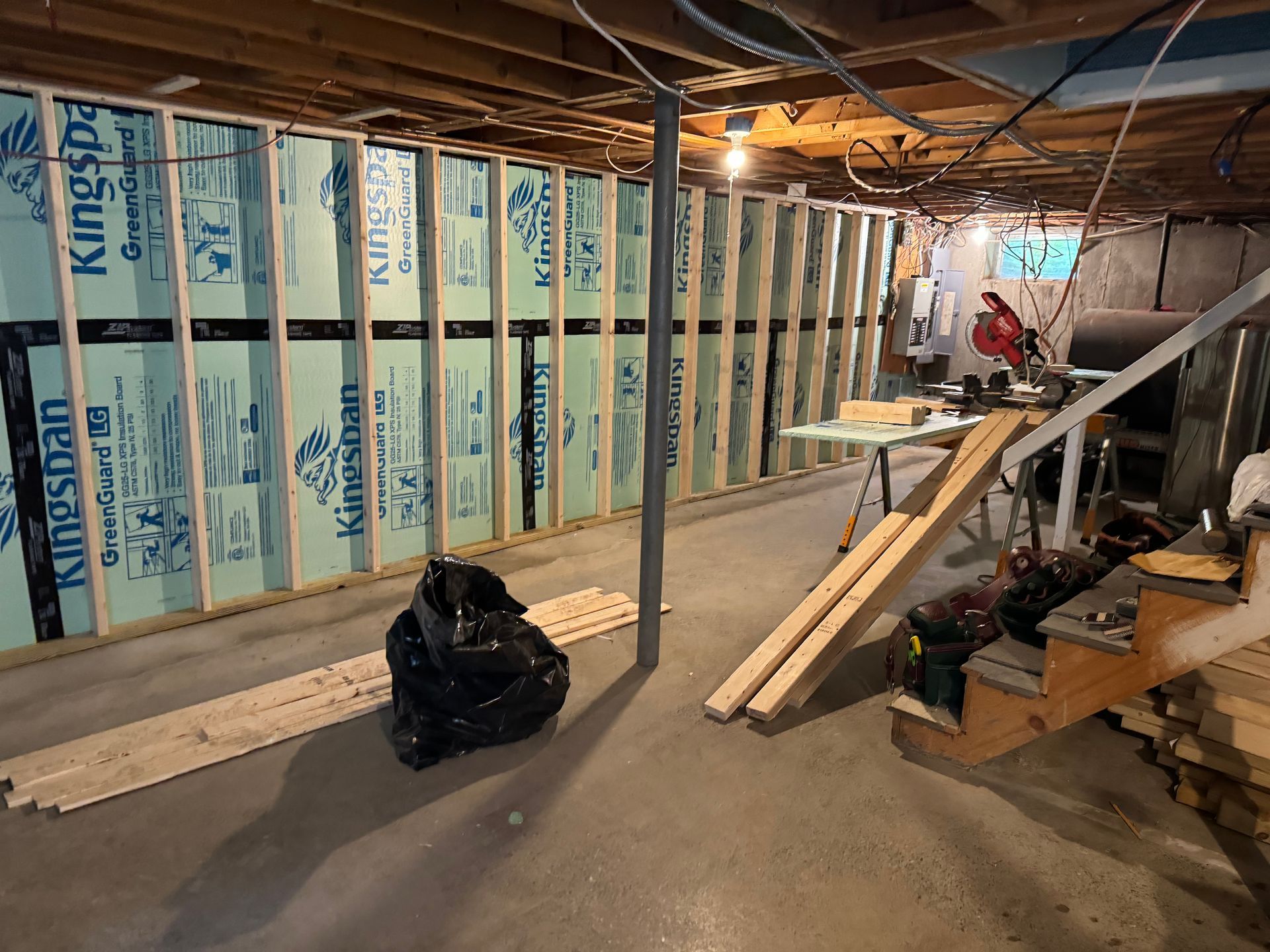 Basement renovation in progress. Blue insulation between wood studs. Saw, stairs, lumber on floor.