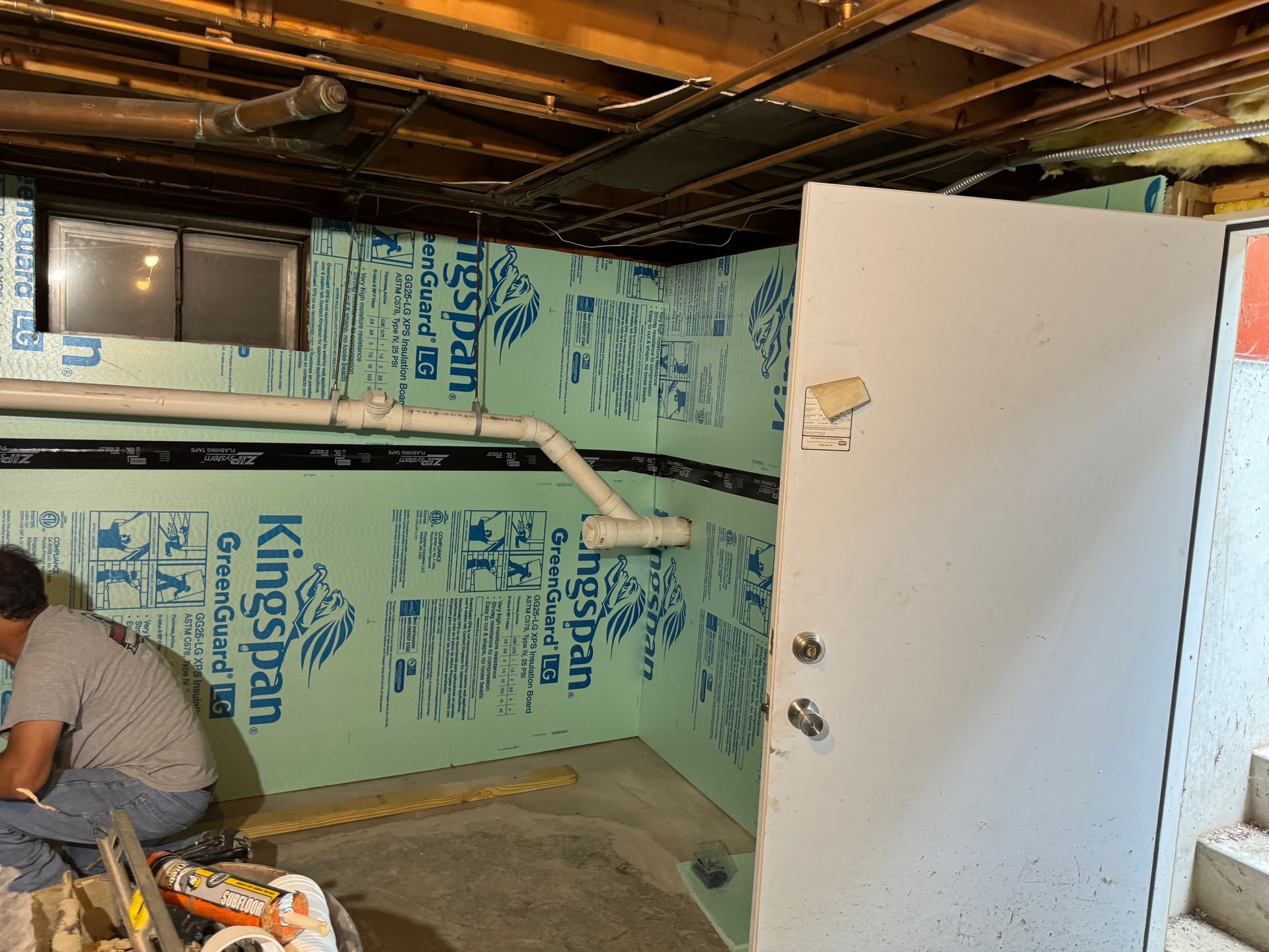 Basement with green insulation on walls, pipes, a door, and a person working.