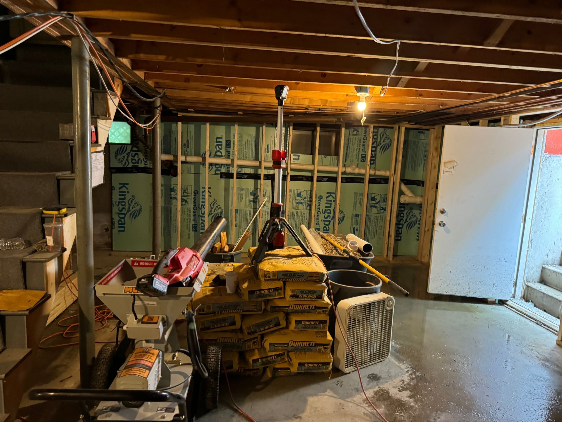 Basement under construction with framing, insulation, tools, cement bags, and wet concrete floor.