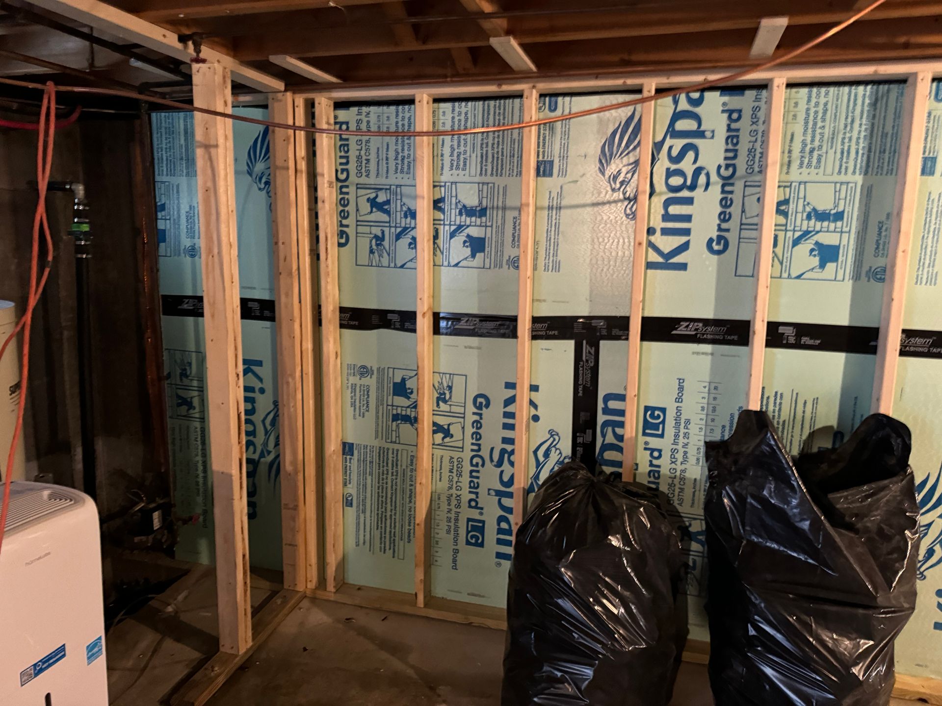Basement with new wooden frame, insulation, and copper pipe; two black bags on the floor.