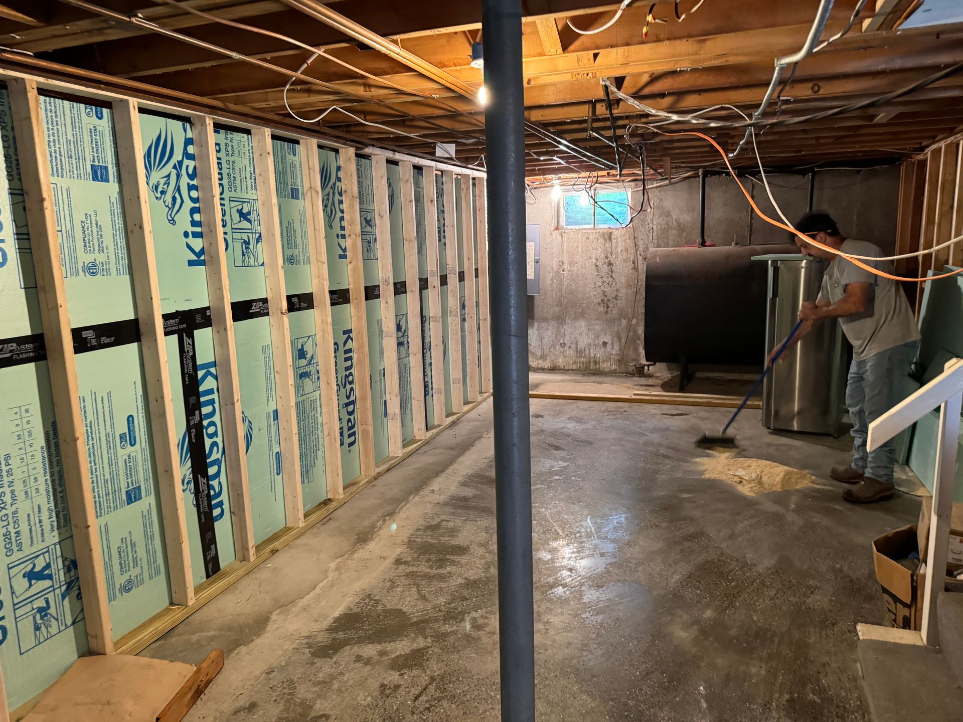 Basement renovation in progress; man sweeping. Exposed studs and insulation on left. Concrete floor, oil tank.