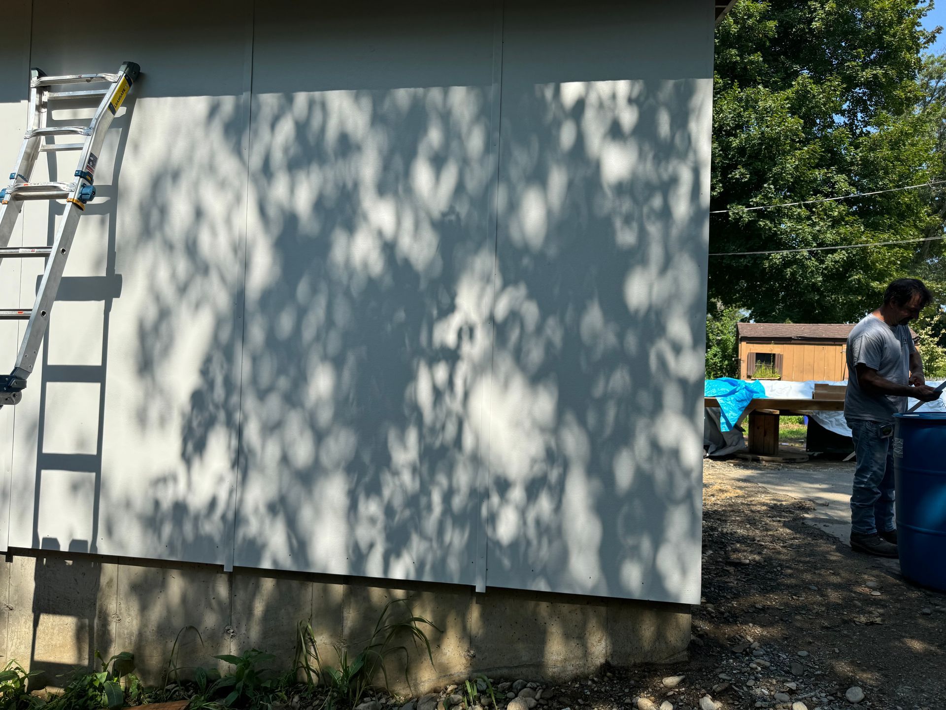 Person painting a light-colored wall, a ladder on the side. Exterior setting with tree shadows and a blue container.