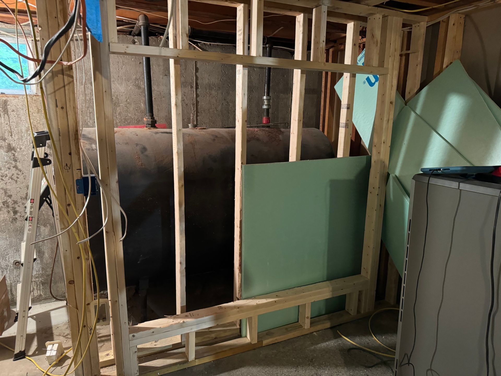 Basement wall framing around an oil tank, with partial green drywall installed.