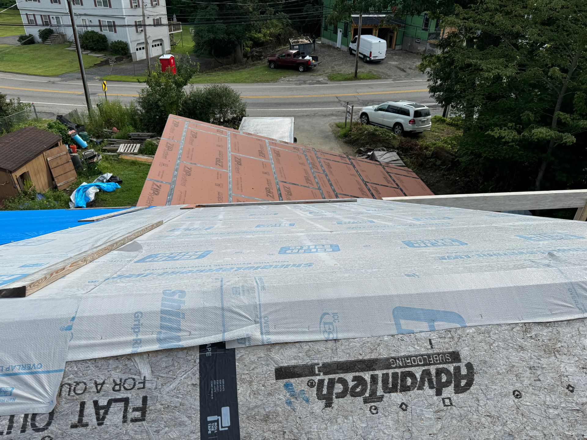 Roofing project with exposed wood panels, tar paper, and partially covered with blue tarp. Street and buildings in background.