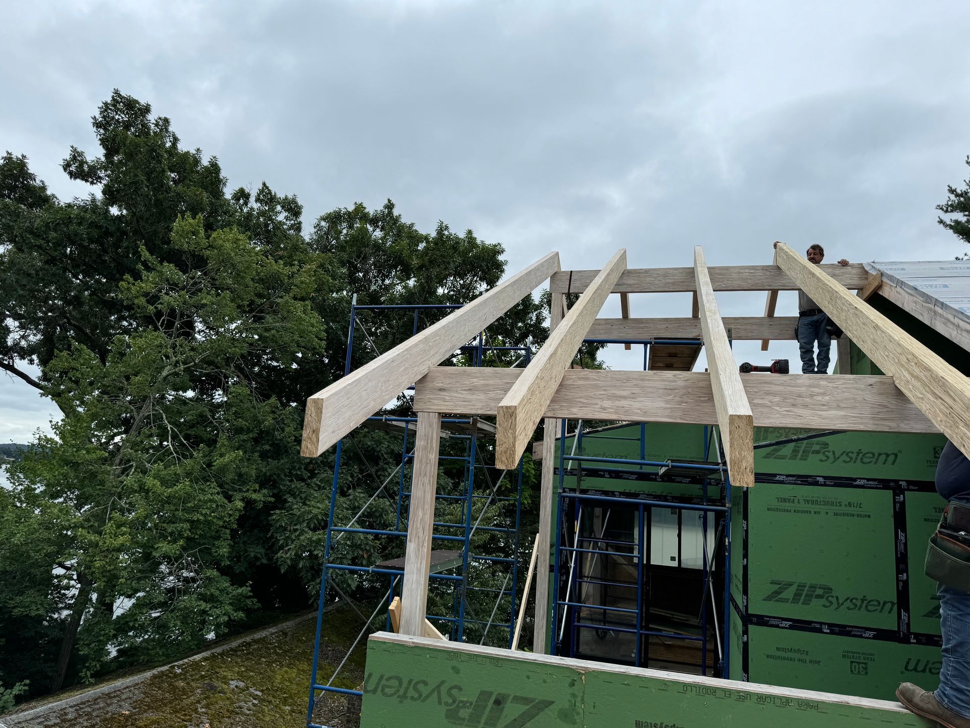 Construction workers building a roof frame. Wood beams on green sheathing with a cloudy sky backdrop.