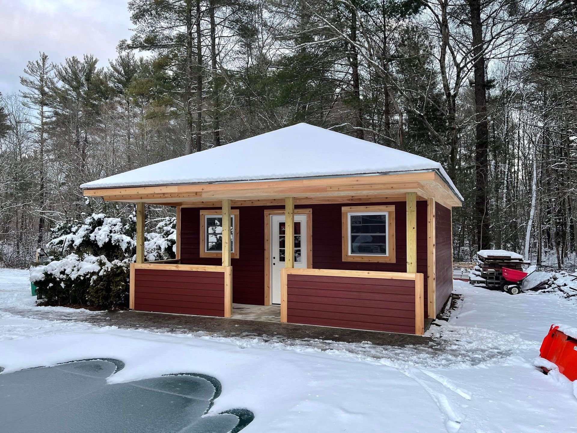 Small red cabin with a porch, covered in snow, surrounded by trees.