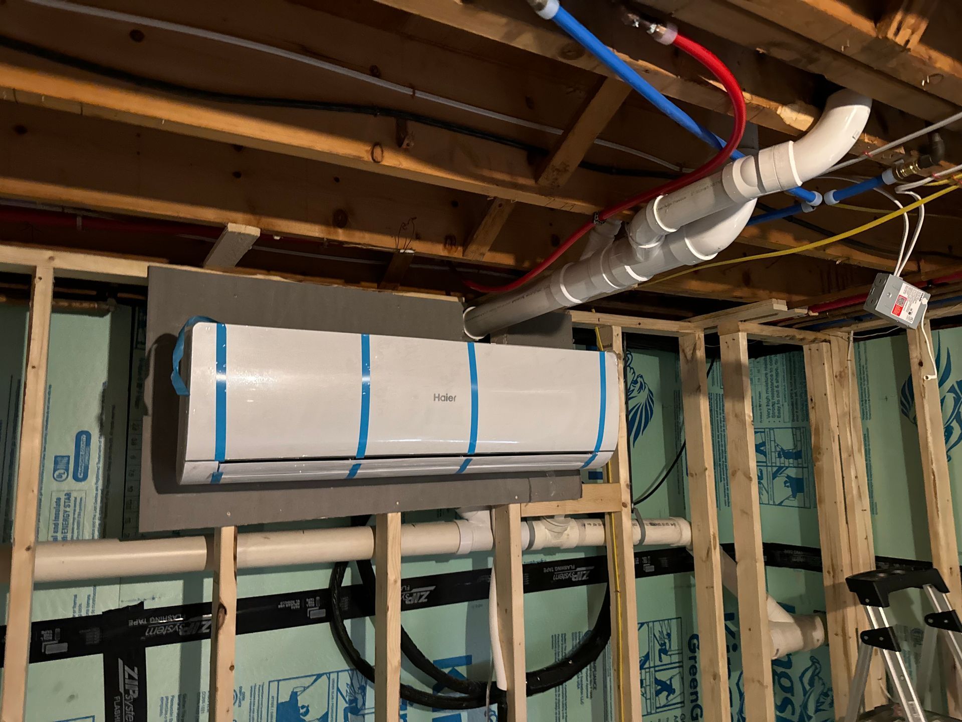 Mini-split AC unit installed on a gray backing board in a basement. Pipes and electrical wiring are visible.
