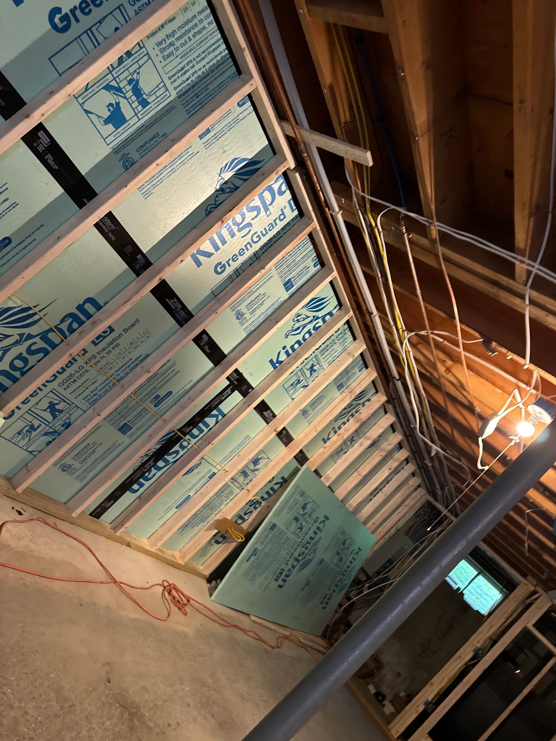 Basement ceiling under construction; blue insulation boards between wooden beams. Wiring and pipes visible.